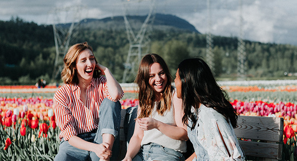 Three women sitting on a bench laughing with a field of flowers and green mountains in the background