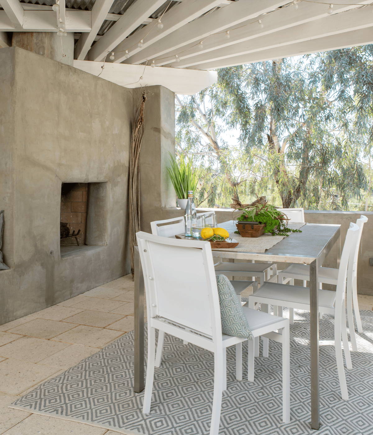 A modern aluminum outdoor dining table sits on a geometric rug under a covered patio with a concrete fireplace in teh background. 