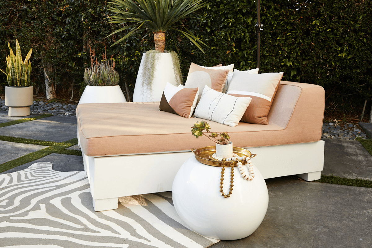 Modern outdoor daybed with blush cushions, accent pillows, a round white side table, and sculptural planters in a styled patio setting.