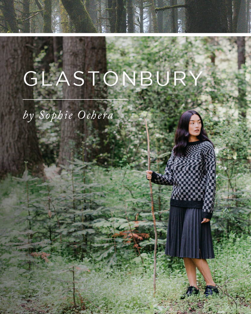 A woman stands in a forest holding a hiking stick. She models a hand knit colorwork sweater with the title GLASTONBURY by Sophie Ochera.