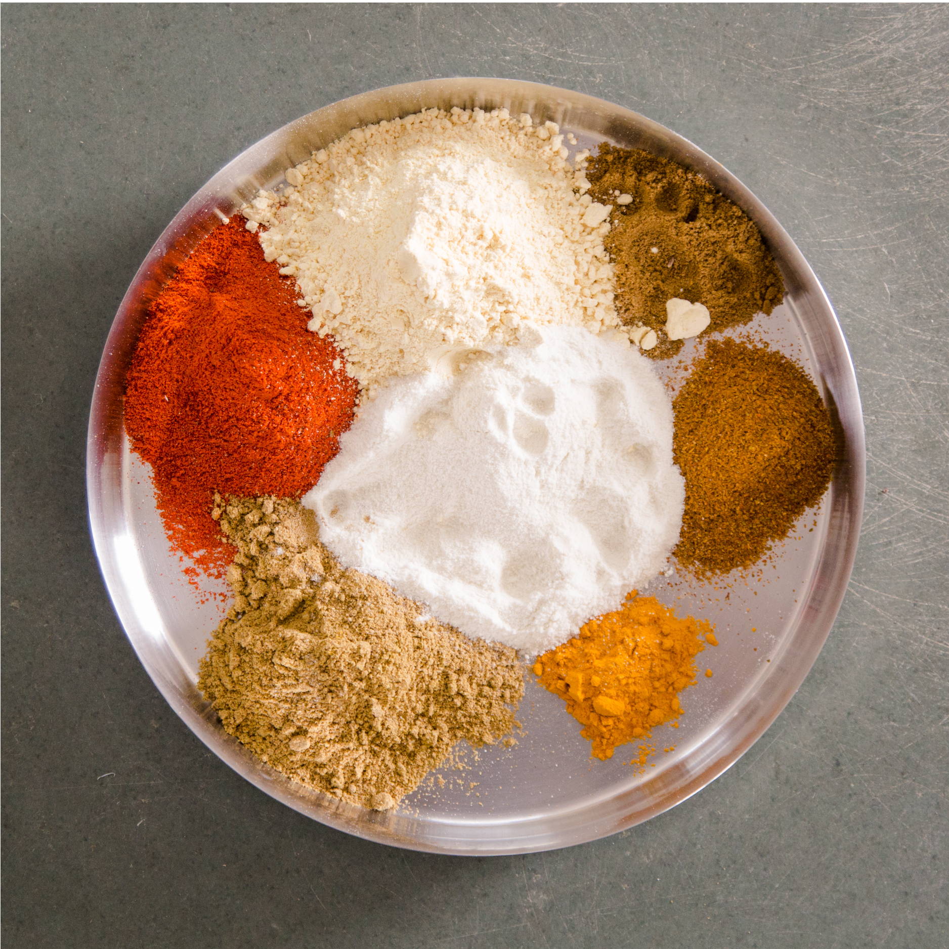 A silver plate sits on a concrete floor. It is holding piles of different Indian spices such as curry, turmeric, and cayenne.