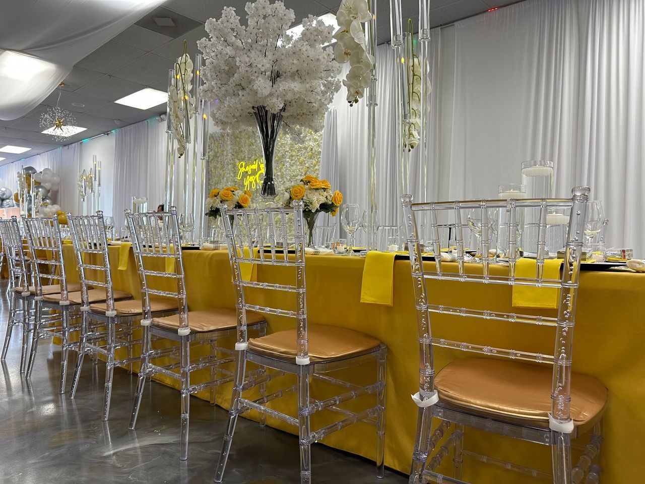 Yellow tablecloth banquet table with clear chiavari chairs and tall white floral centerpiece
