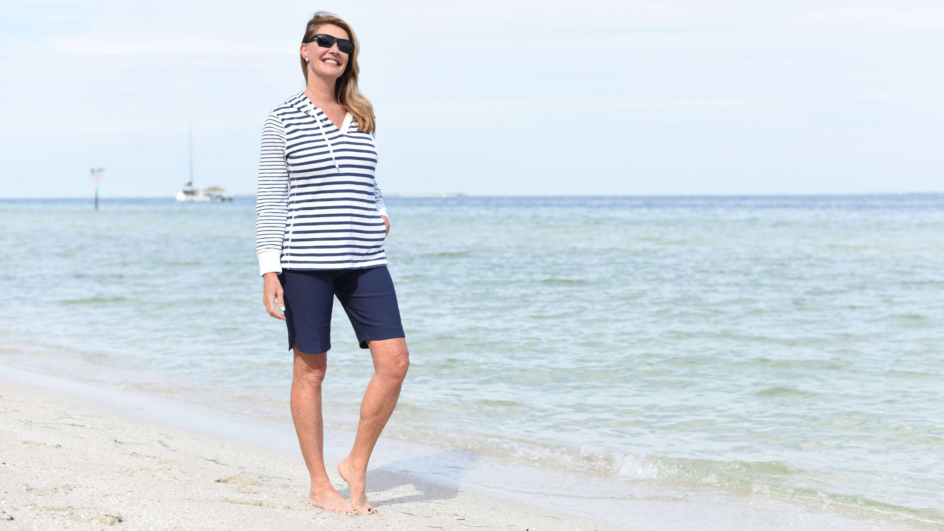 A smiling blonde woman wearing sunglasses, a striped hoodie, and bermuda shorts stands on a beach.