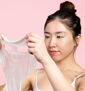Woman examining a translucent skincare mask against a pink background.