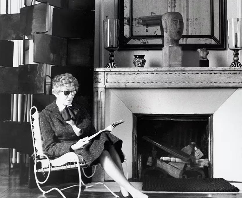 Older Eileen Gray sitting on chair in front of fireplace.