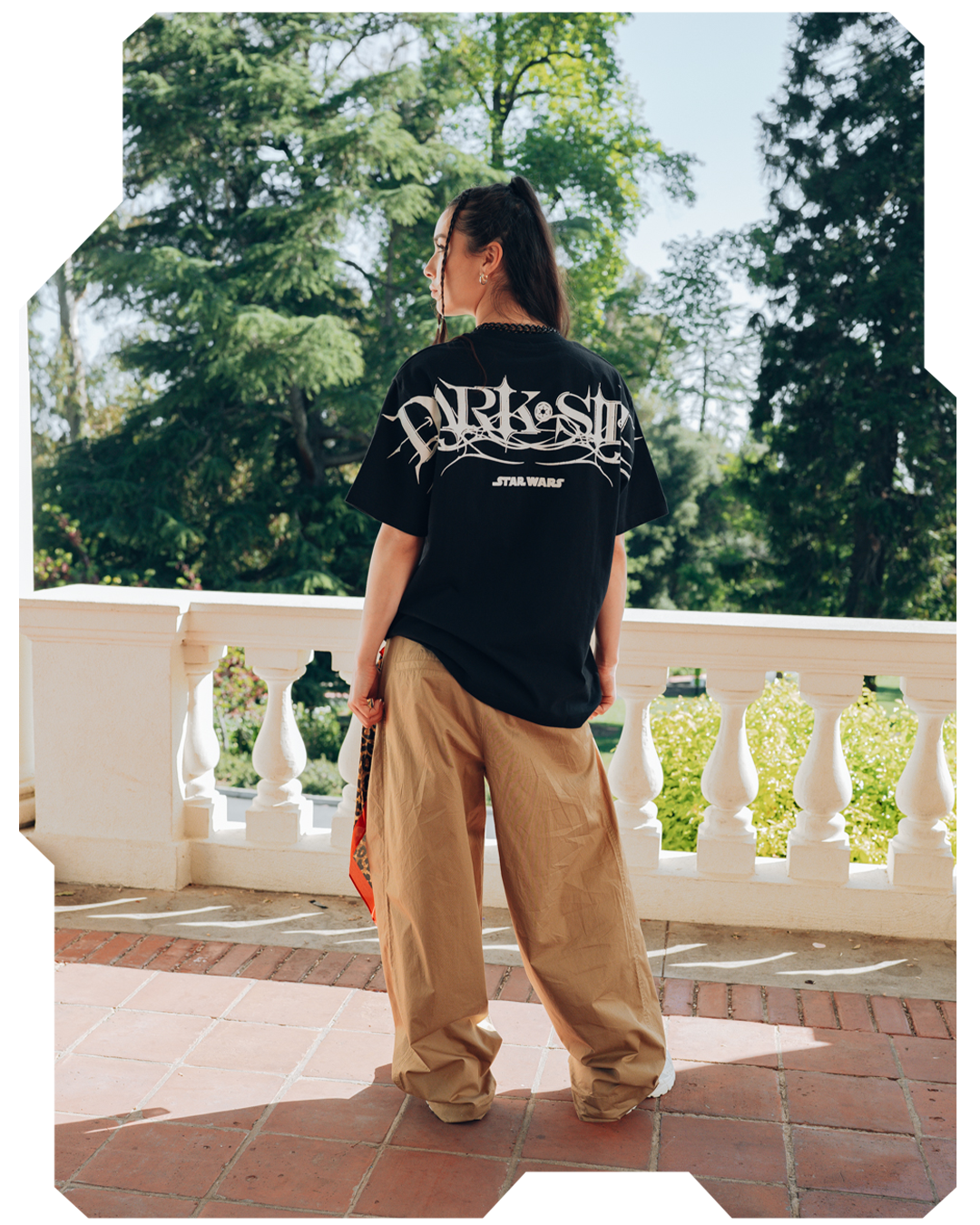 Person wearing a black t-shirt and beige pants, standing outdoors near a railing.