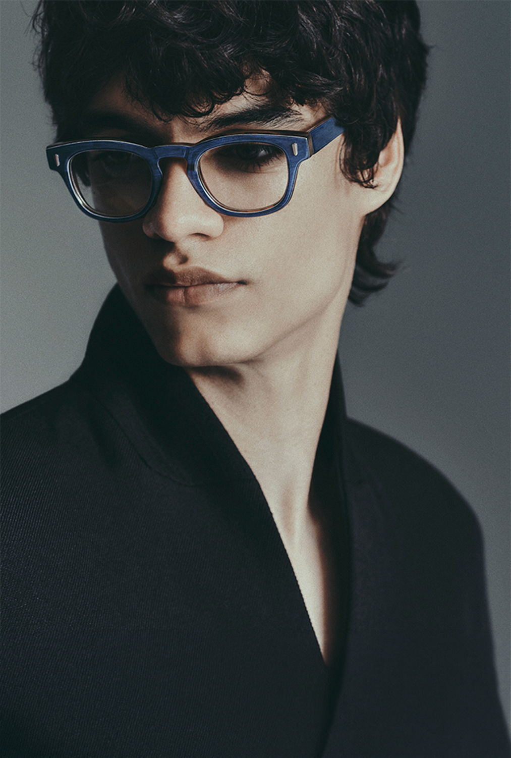 Young man with curly hair and blue glasses, wearing a black jacket.