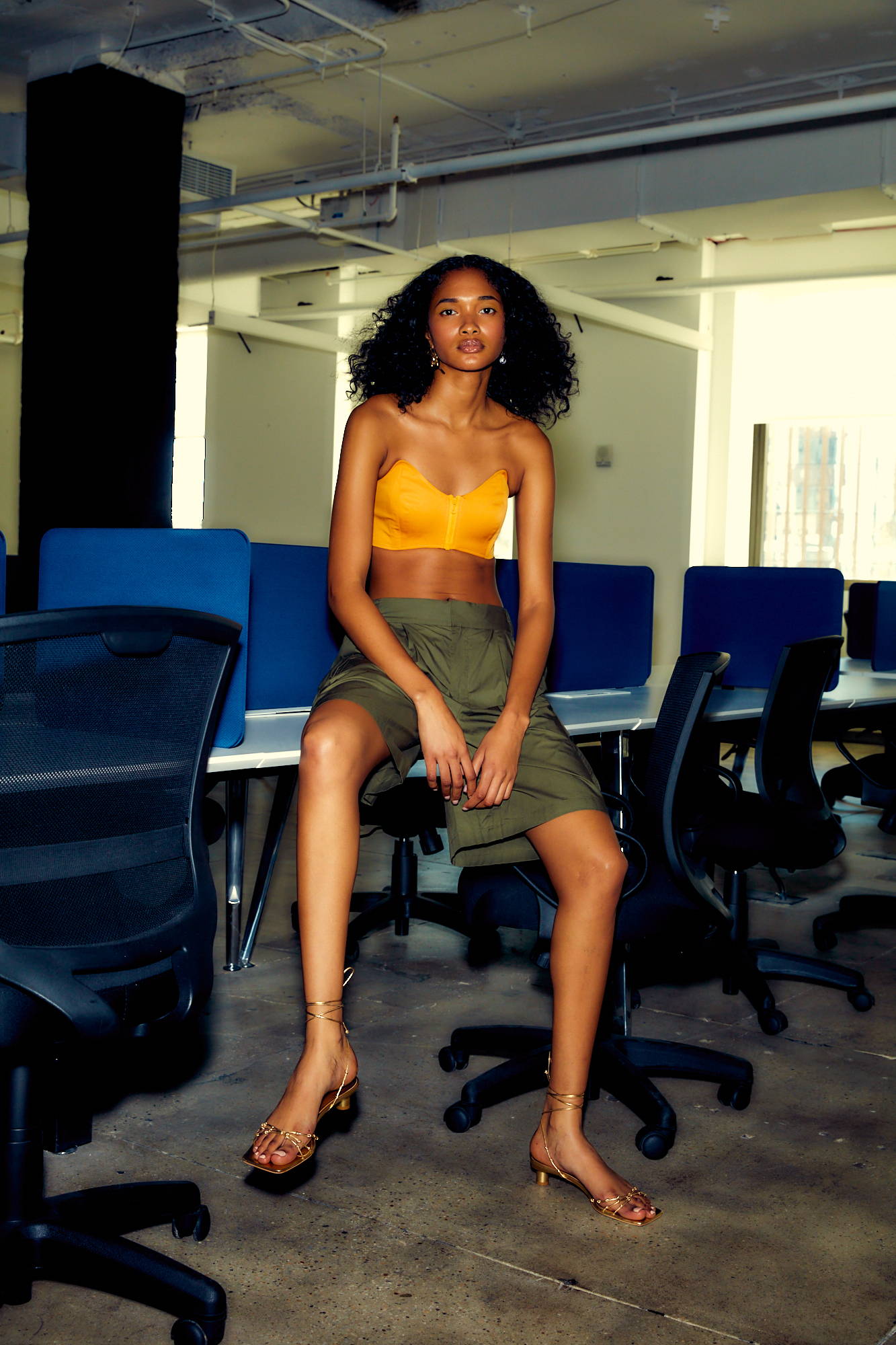Woman in an office, wearing a yellow top and skirt, sitting on a chair.