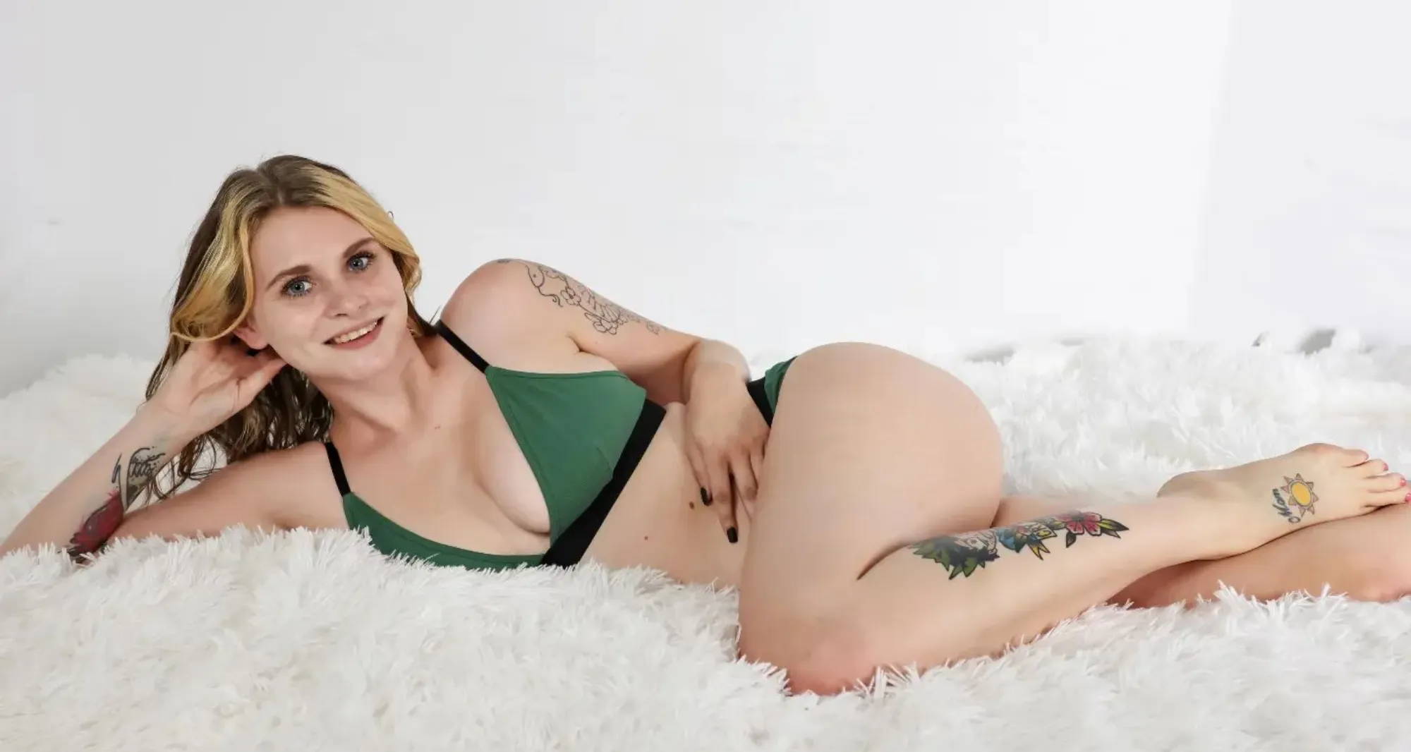 blonde woman with tattoos laying on one side on a white fluffy rug wearing a green triangle bra and undies set