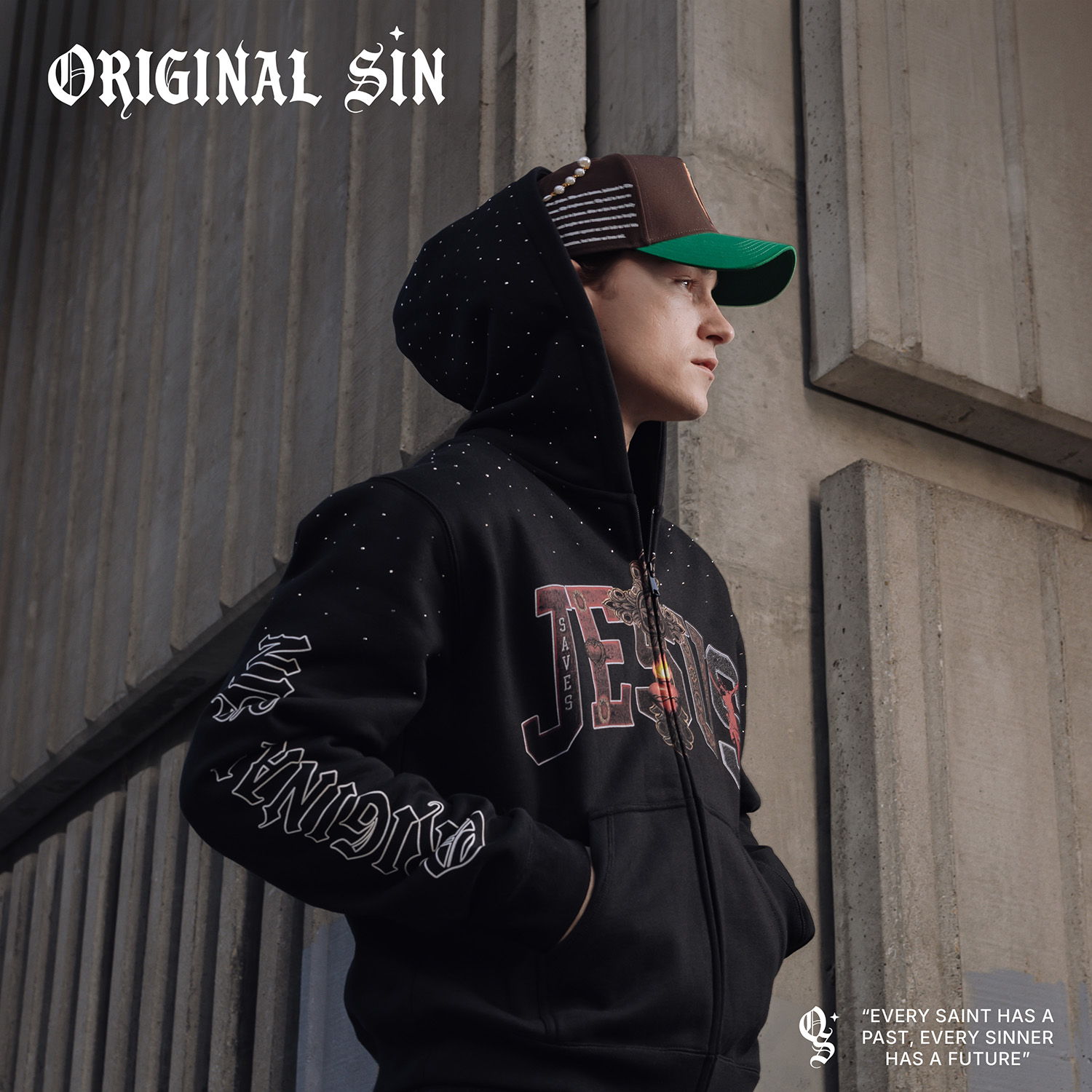 Person in a black hoodie and cap standing against a textured wall.