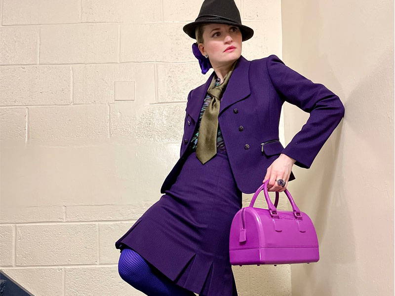 A woman wearing a purple suit and skirt with a olive necktie and pink handbag