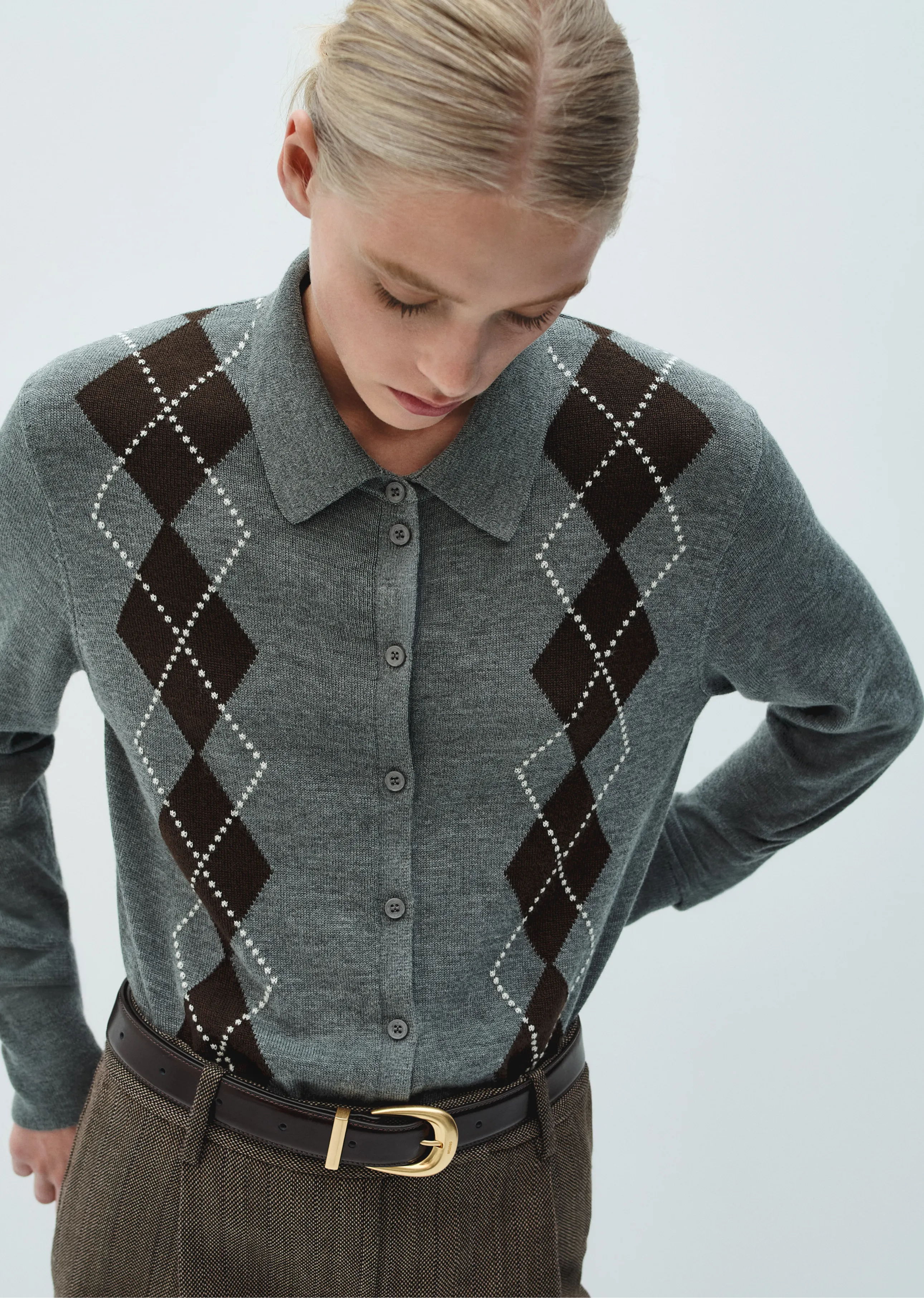 Woman in a gray argyle sweater with a collar, wearing brown pants.