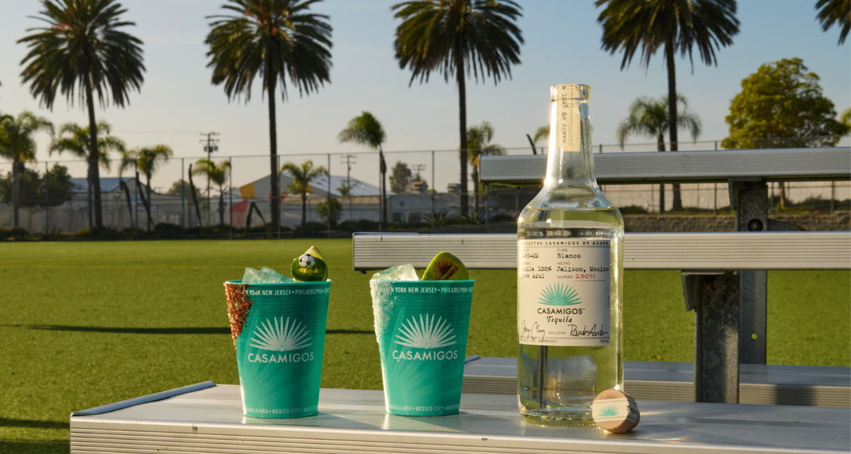 Two teal cups and a bottle of Casamigos tequila on a table with a soccer pitch and palm trees in the background.