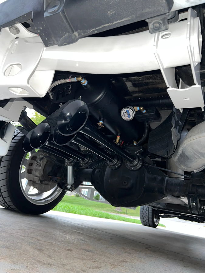 Underside of Cade's GMC Sierra 2500HD showing four black Conductor's Special 244 Nightmare Edition train horn bells cylindrical air tank blue airlines pressure gauge mounted between white frame rails behind rear axle photo 2