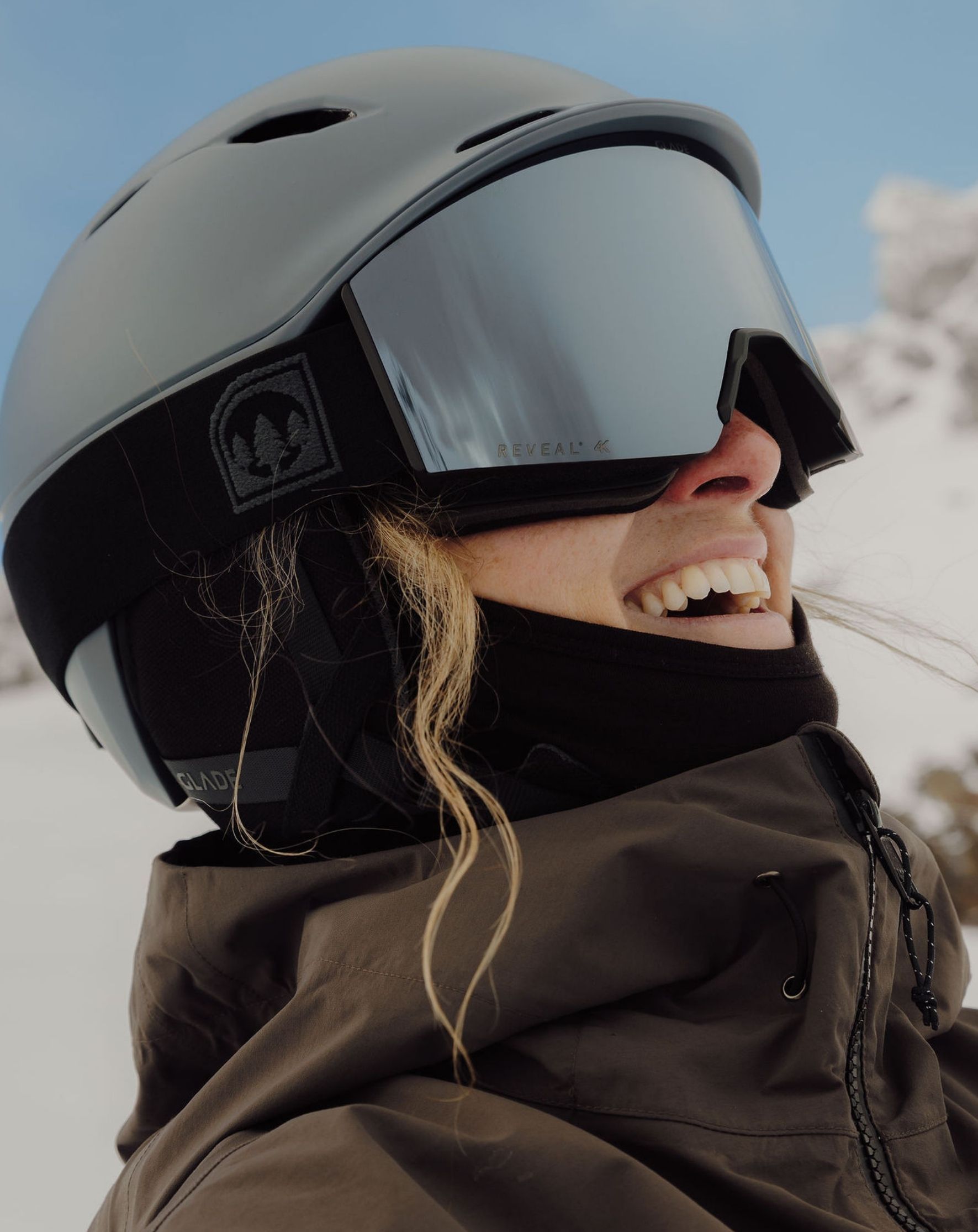 Person smiling while wearing a ski helmet and goggles in a snowy environment.