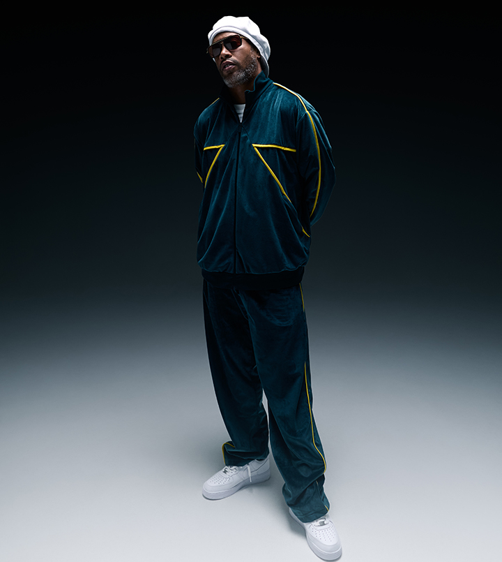 A person in a tracksuit and hat, squatting against a dark background.