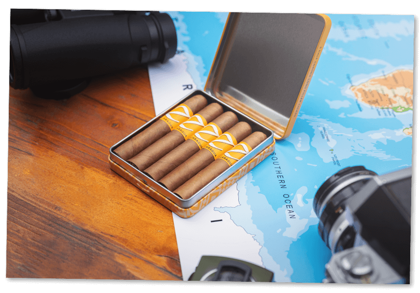 Five pre-cut Zino Nicaragua Half Corona cigars in their opened tin, which is placed on top of a world map between a camera and a pair of binoculars. 