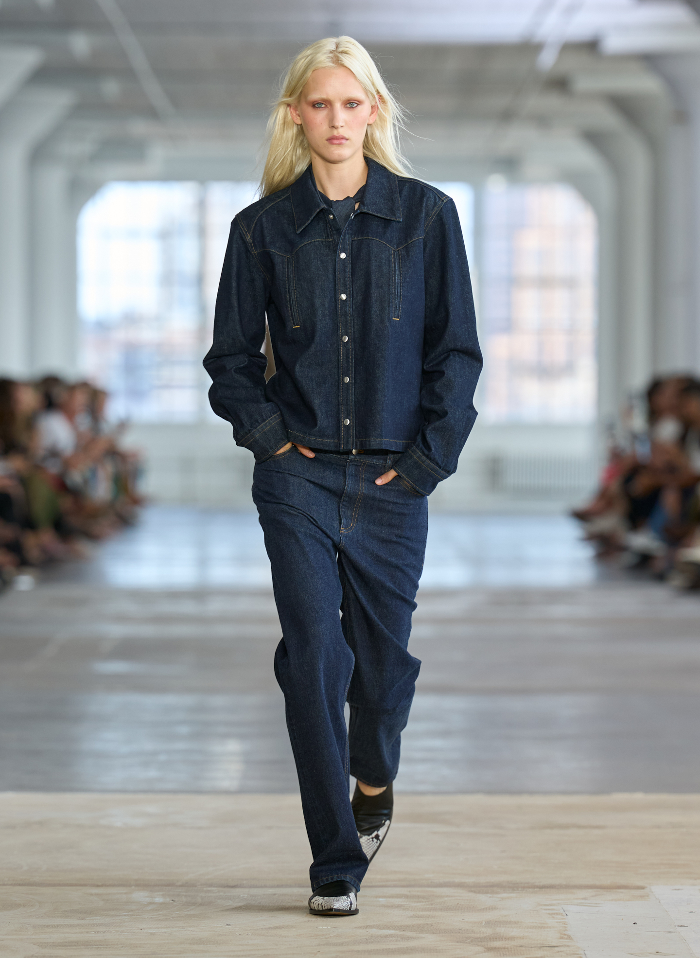 Model walking down runway wearing denim outfit in a spacious venue.