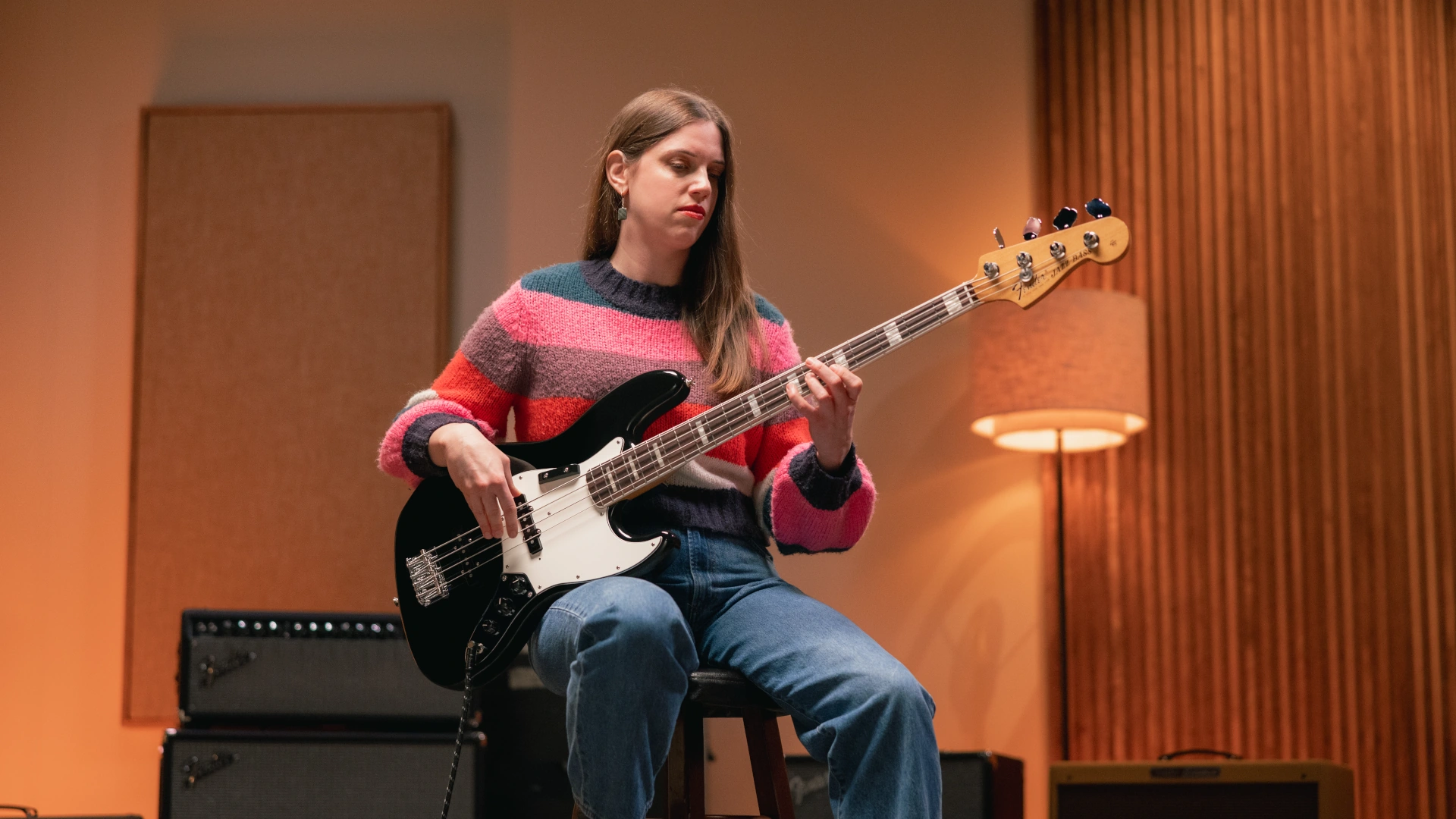 Woman playing a black Fender Vintera III Early '70s Jazz Bass guitar while sitting on a stool.