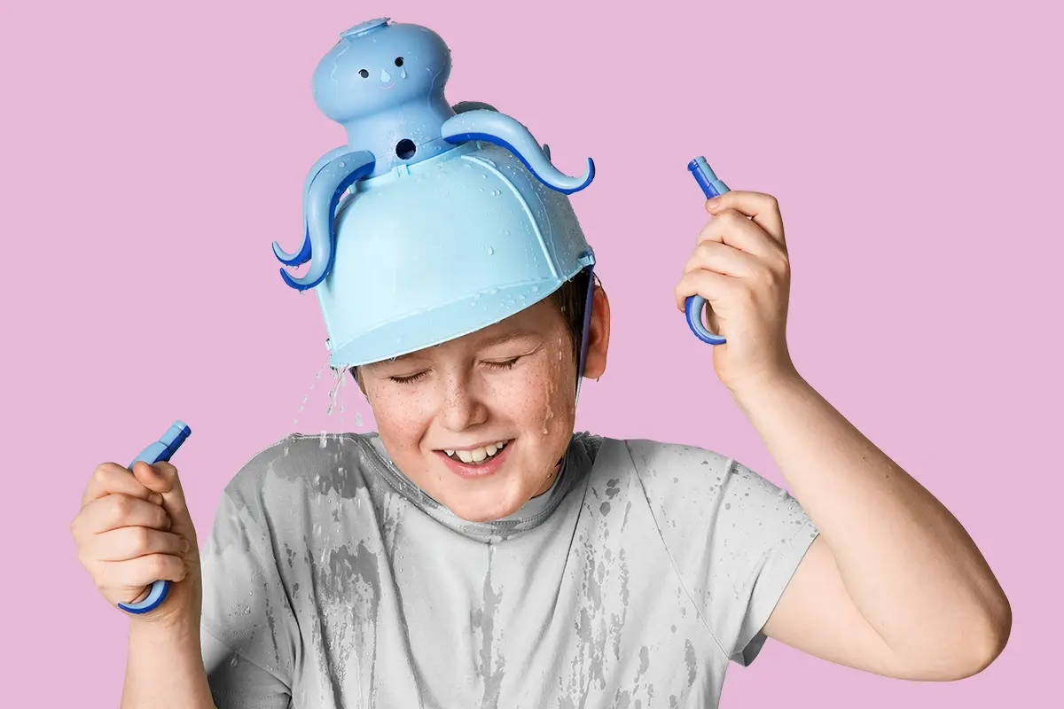 Child wearing a blue octopus water hat playing with water crayons, set against a pink background