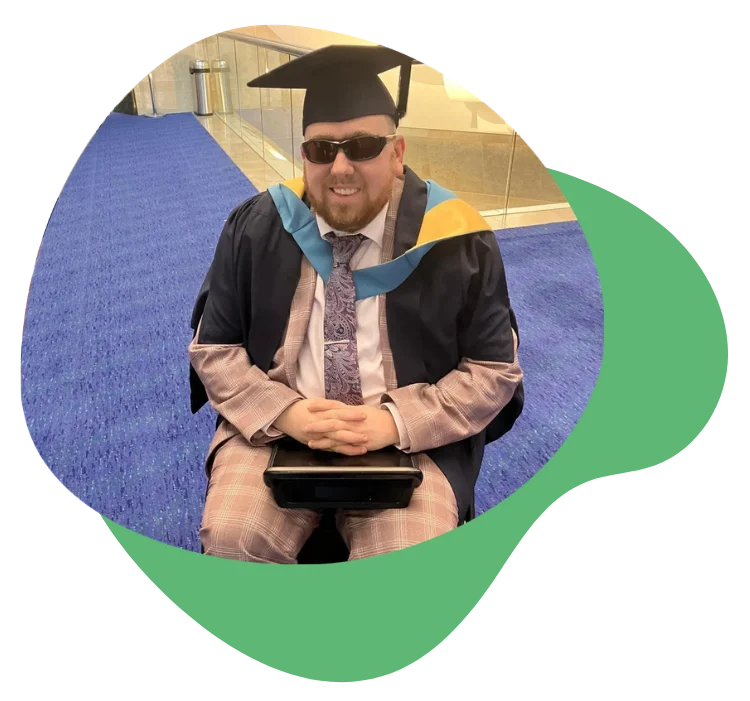 Person in graduation attire seated in wheelchair, wearing sunglasses, smiling.