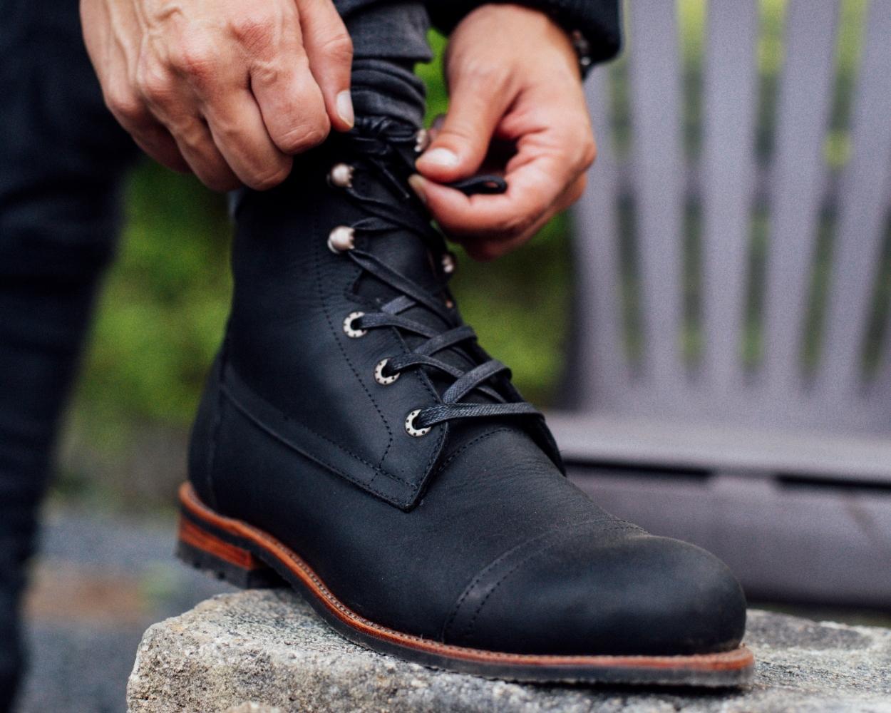 A man tying the laces of his Adelante black full-grain leather wide width boots while stepping on a stone outside.