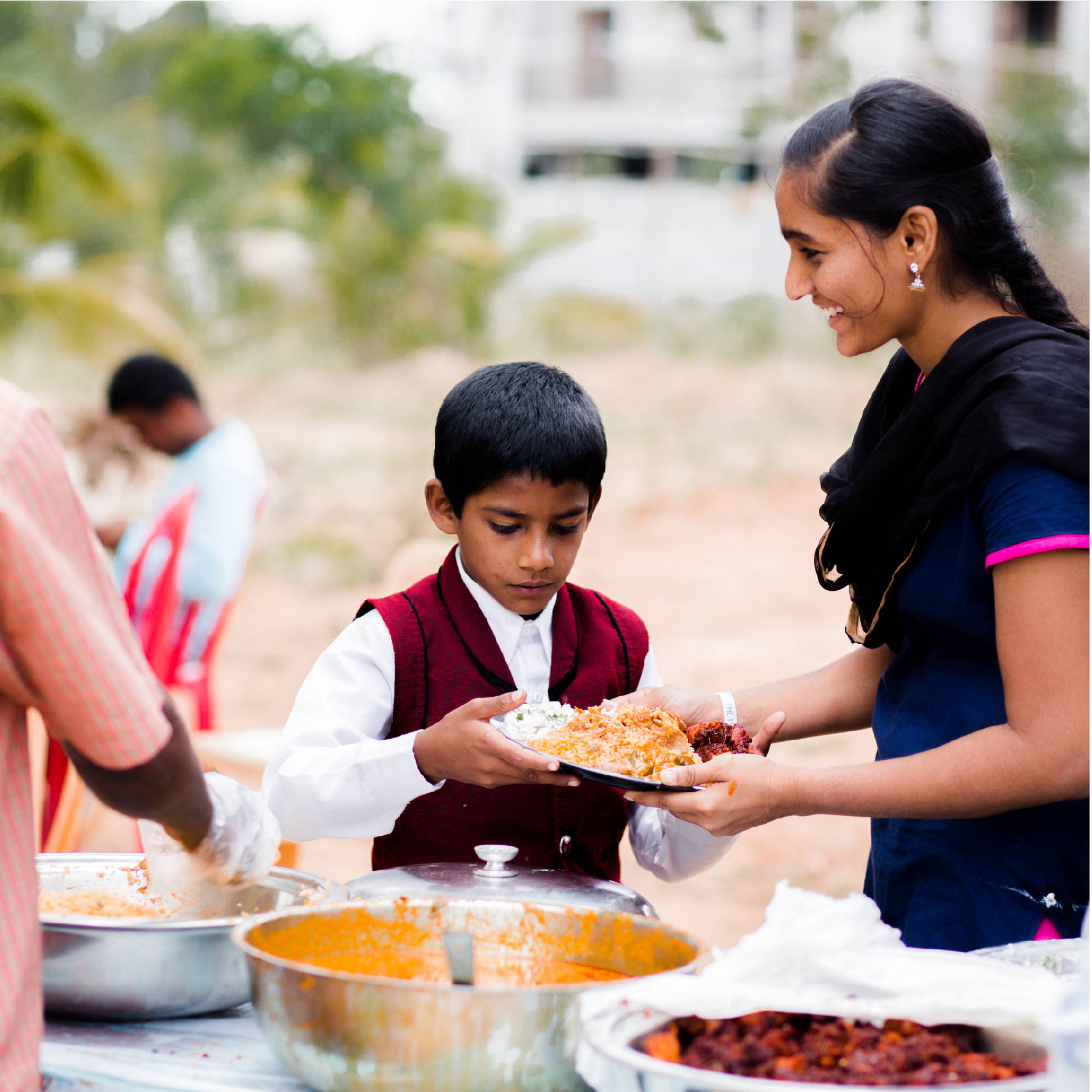A young boy in red is served a plate of Indian food by a smiling teenage girl at Violet's Children's Home. 