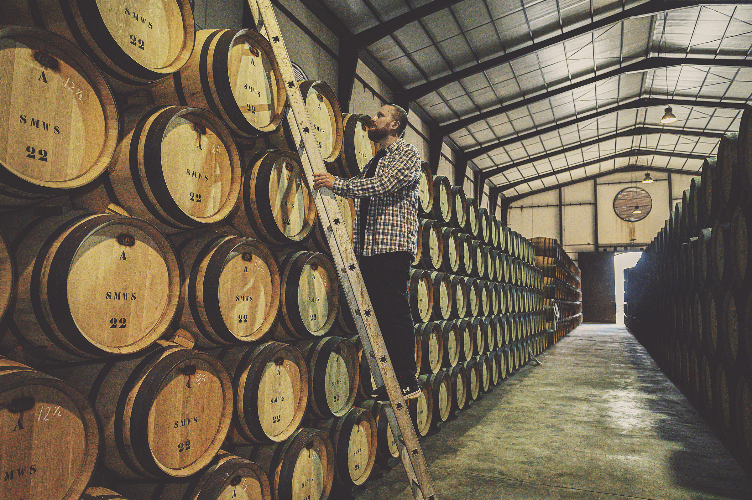 Person climbing a ladder near stacked wooden barrels in a warehouse.