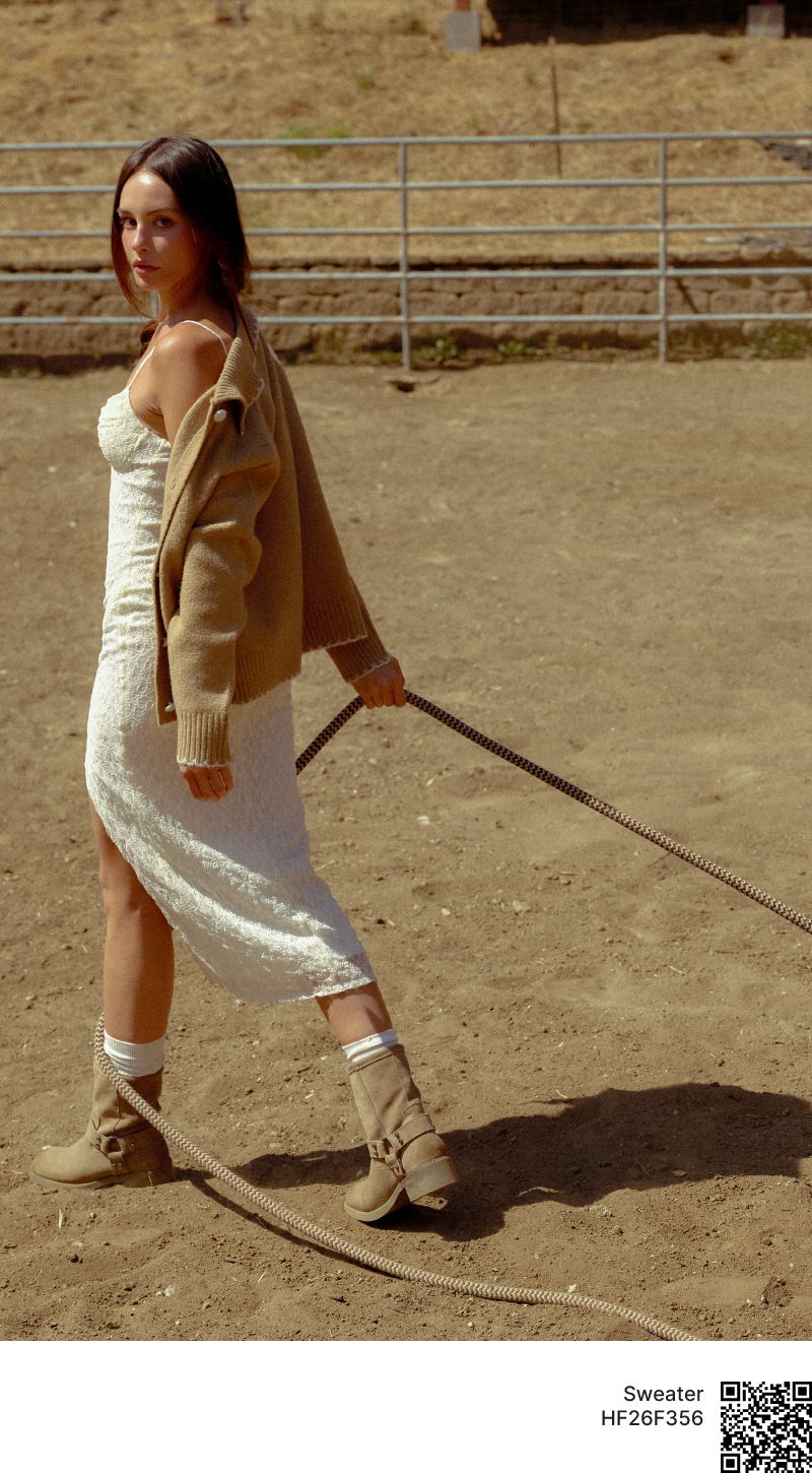 Woman in a white dress and beige jacket, walking in a dusty arena.