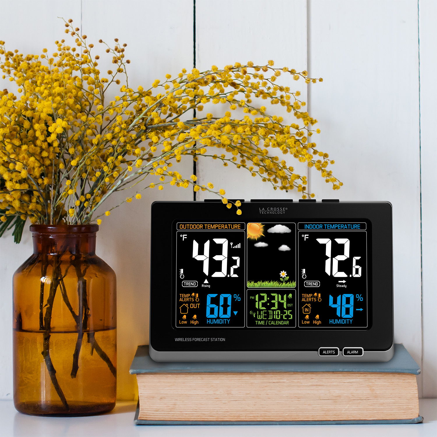 Weather station on books beside a vase of yellow flowers.