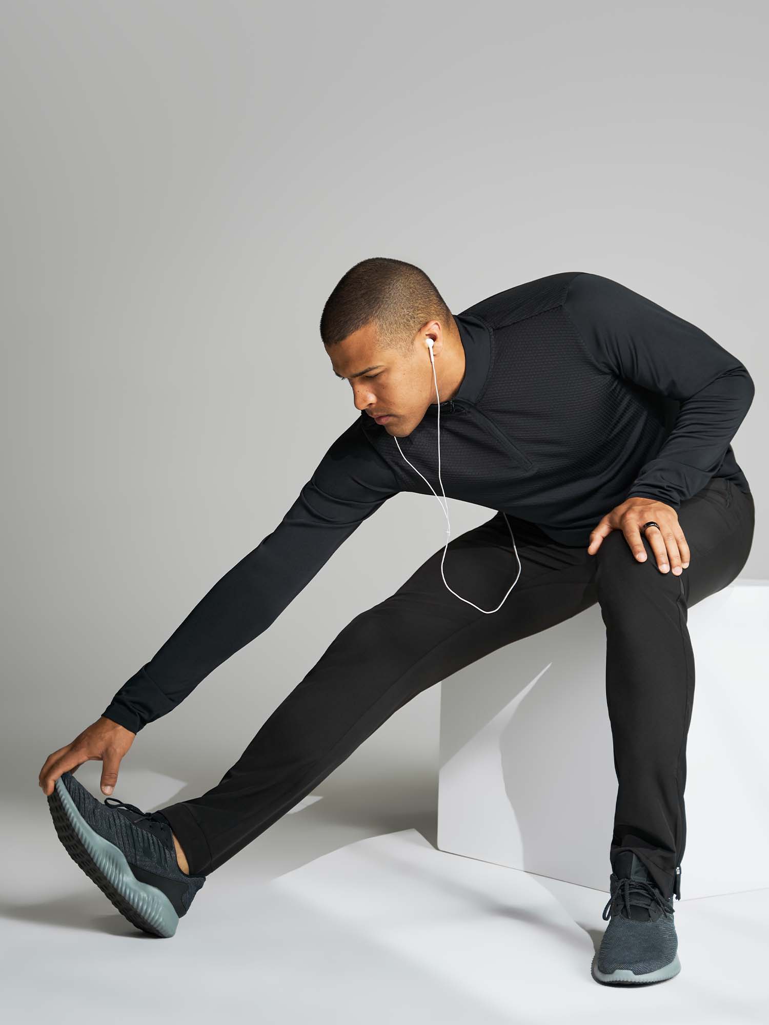 Tall man sitting on a block stretching his leg while wearing a black athletic track suit and running shoes