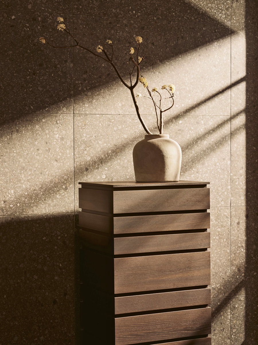 A vase with branches sits on a wooden Lema chest in soft light.