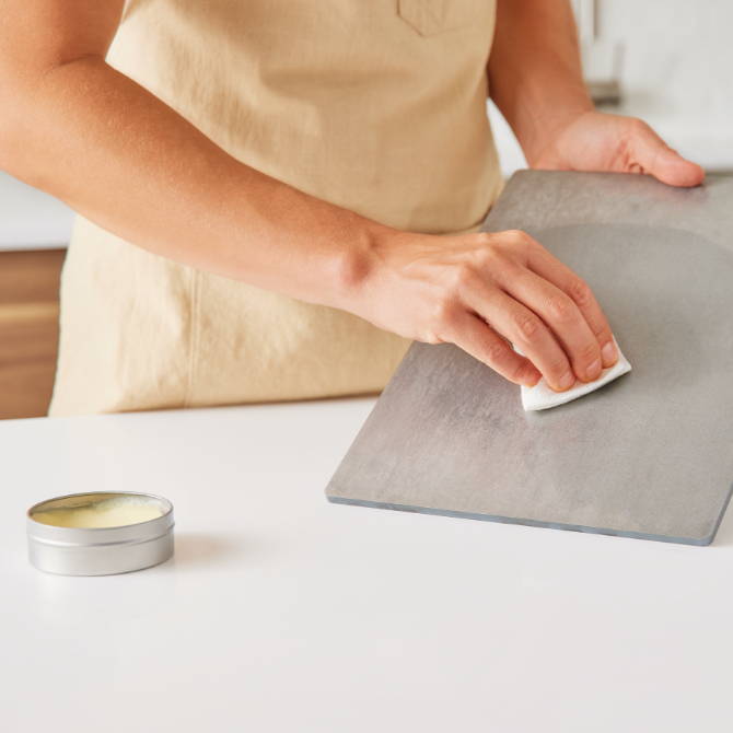 A chef uses a paper towel to wipe a thin layer of seasoning wax on a Misen Oven Steel. A tin of Misen Carbon Steel Seasoning Wax is visible.