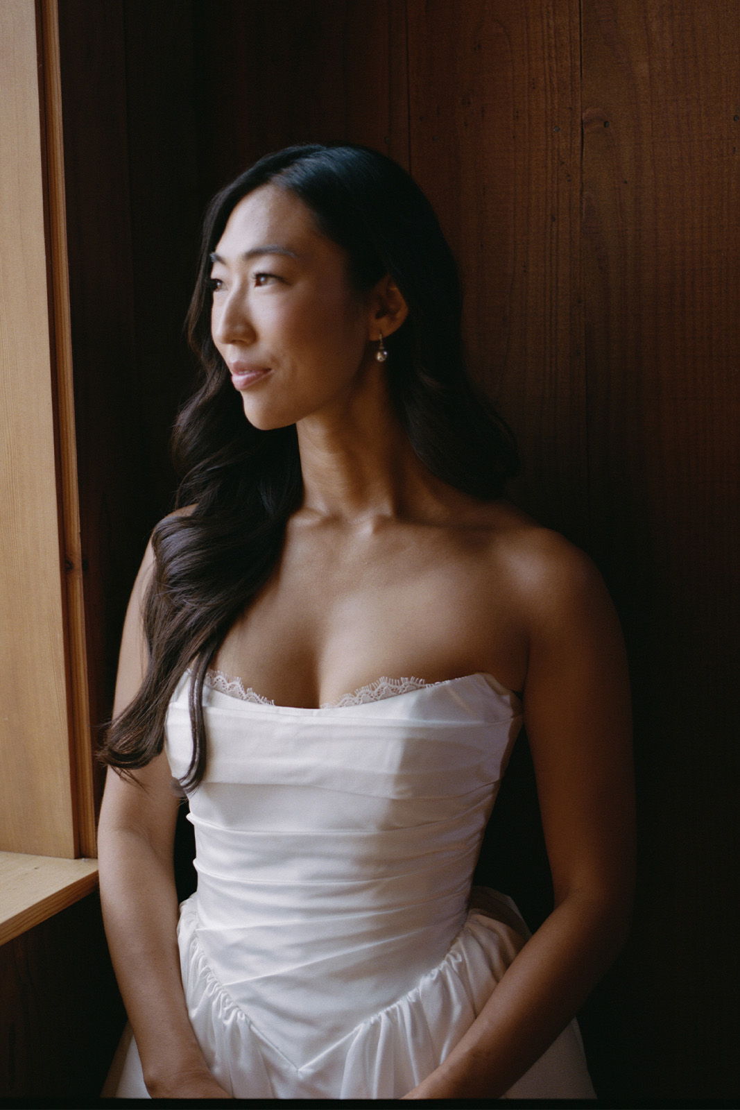 Woman in a wedding dress, gazing out a window with a thoughtful expression.