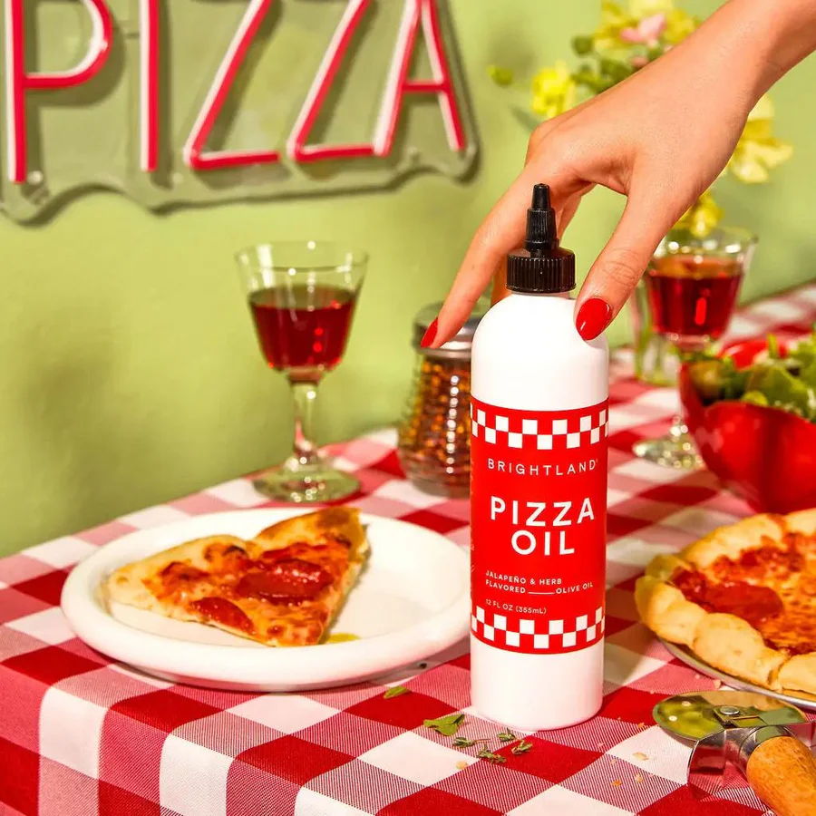 Hand holding pizza oil bottle near pizza slice on a checkered tablecloth.