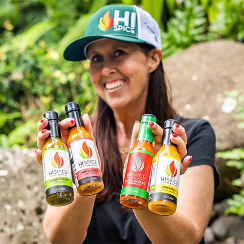 Person holding four bottles of sauce, smiling outdoors in a hat.