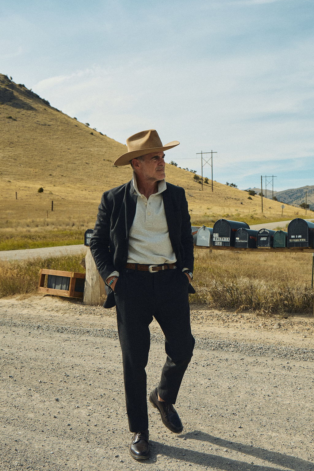 Michael Kelly walking down a gravel road wearing a vintage Stetson, the Rivay Shelby Italian Wool Suit in Charcoal Herringbone, the Torino Cotton Cashmere Fleece Polo and the Alden Plain Toe Blucher 990 in Color 8 Shell Cordovan