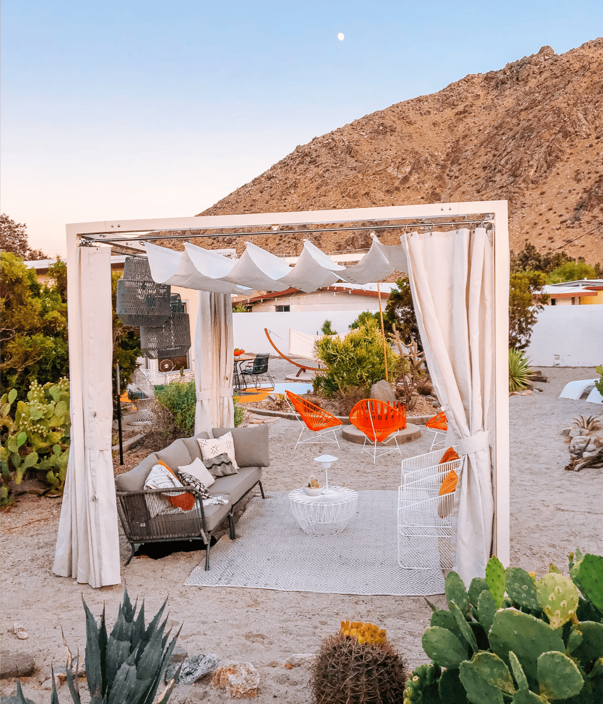 A shaded cabana in a desert patio with lounge sofa and white wire chairs. 