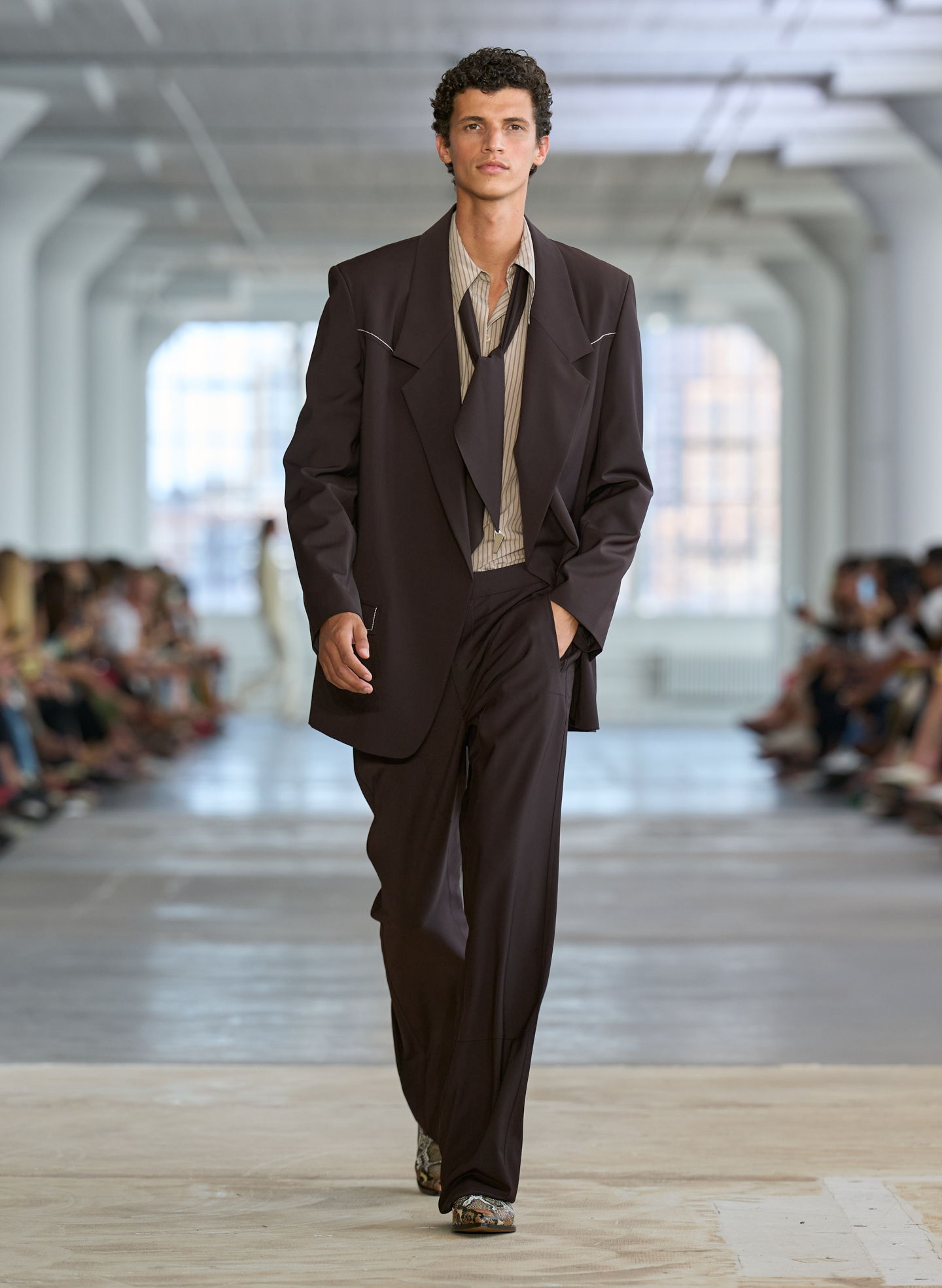 A male model walks the runway in oversized dark suit attire.