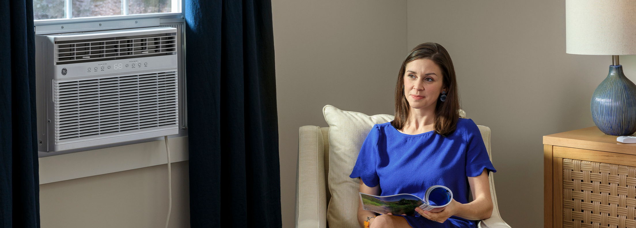 Woman in blue dress sitting with a book near an air conditioner.