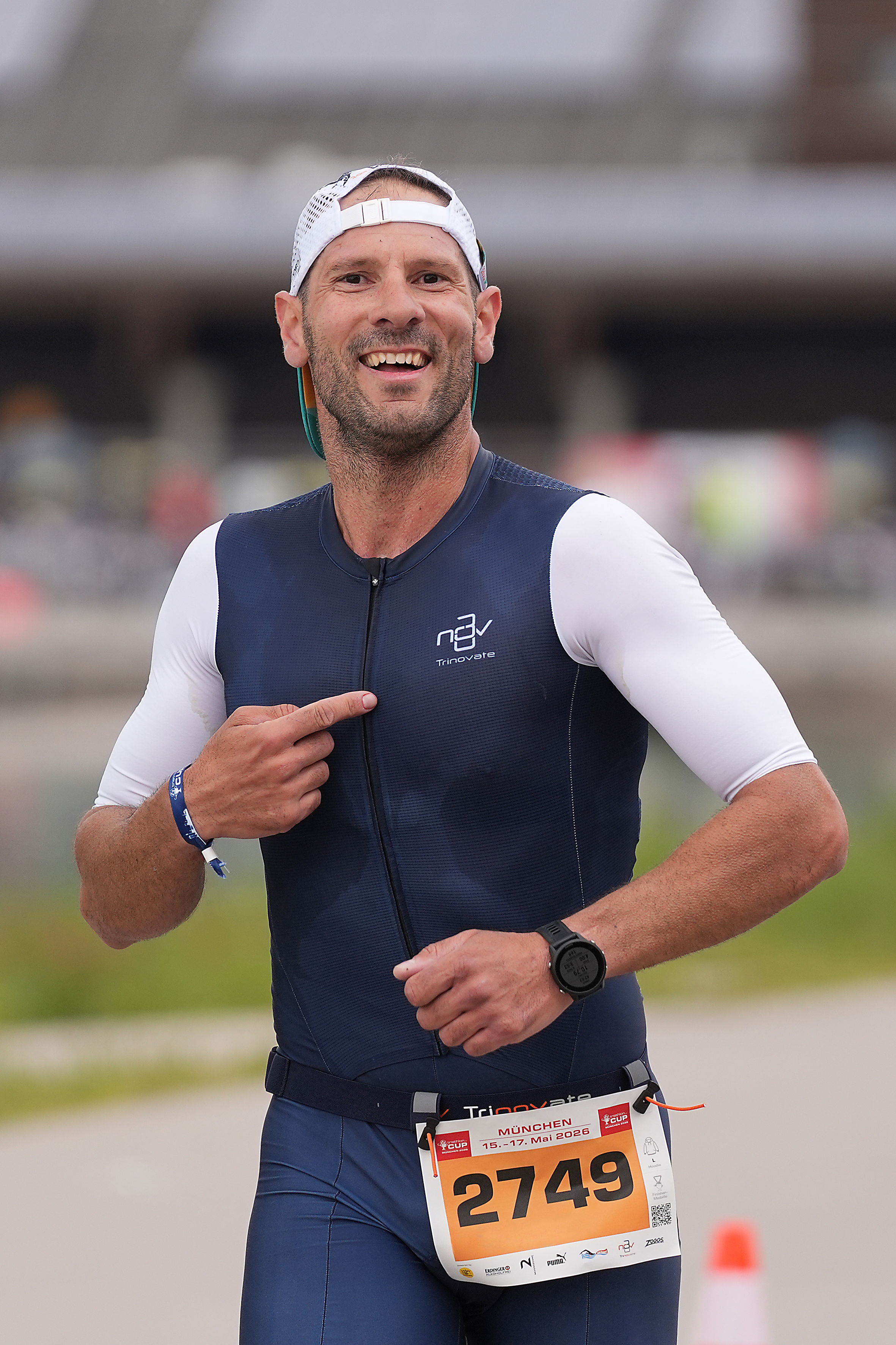 Ein Triathlet im blau-weißen Trinovate Einteiler. Er lächelt in die Kamera und zeigt auf das Logo auf seiner Brust