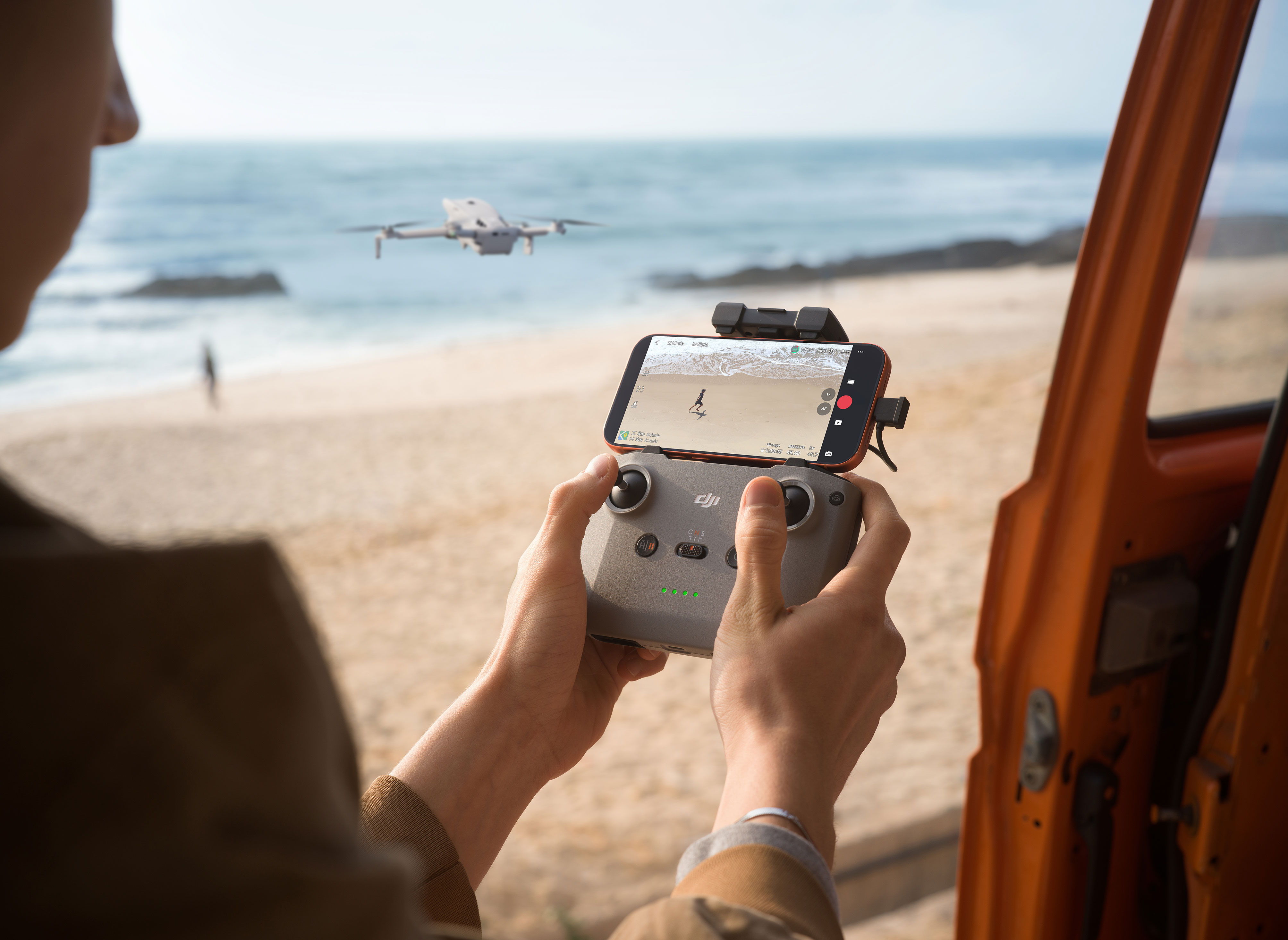 Person controlling a DJI Lito Series drone on a beach with a smartphone connected to the DJI RC-N3 controller.