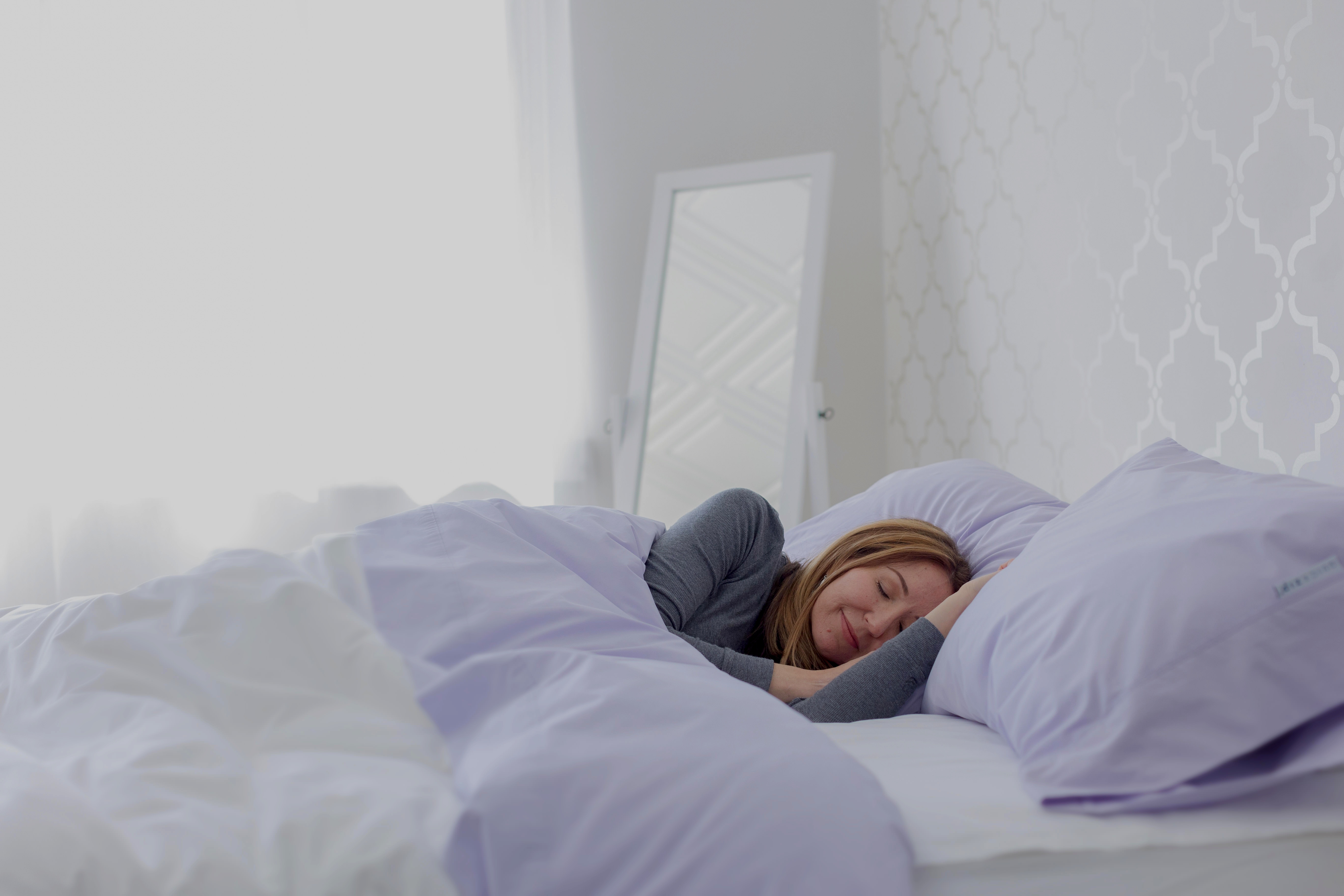Woman sleeping peacefully on white and lavender sheets. the room decor is white ant there is a mirror and window in the background.