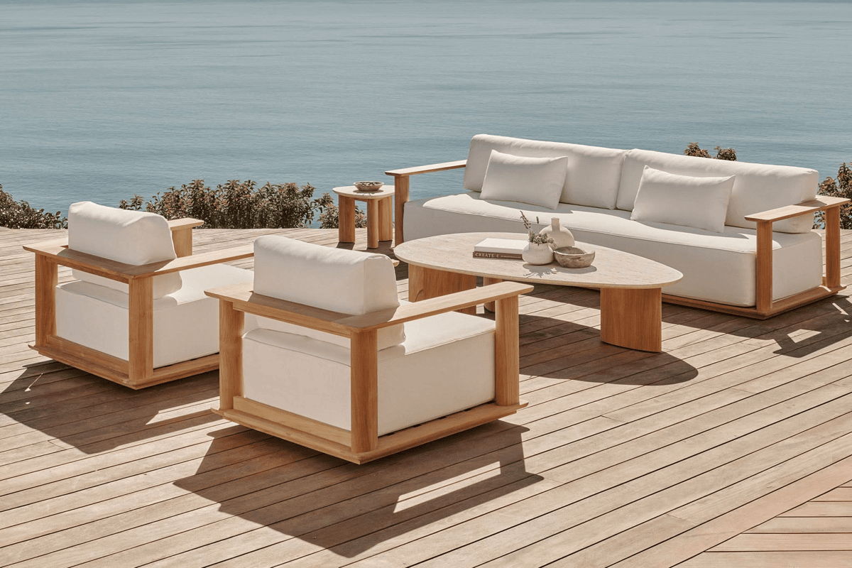 Teak-frame outdoor sofa and armchairs with white cushions on a wood deck facing an ocean view, with an oval coffee table.