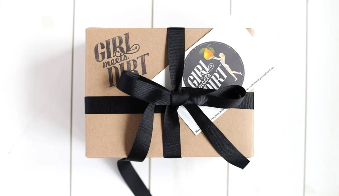 Small craft paper box with Girl Meets Dirt logo stamped on top left corner, wrapped with a black satin bow. Girl Meets Dirt postcard is tucked into the ribbon.