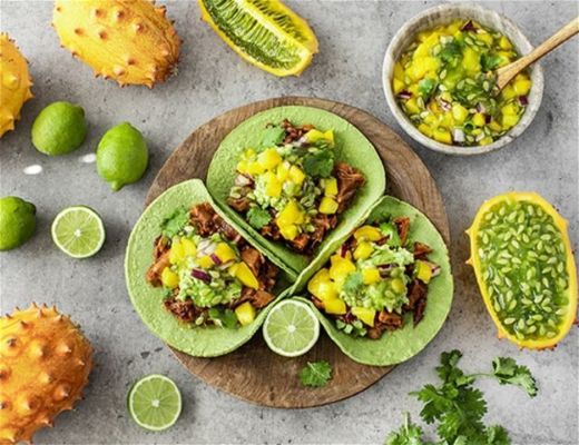 Three tacos with toppings, lime, and tropical fruits on a gray surface.
