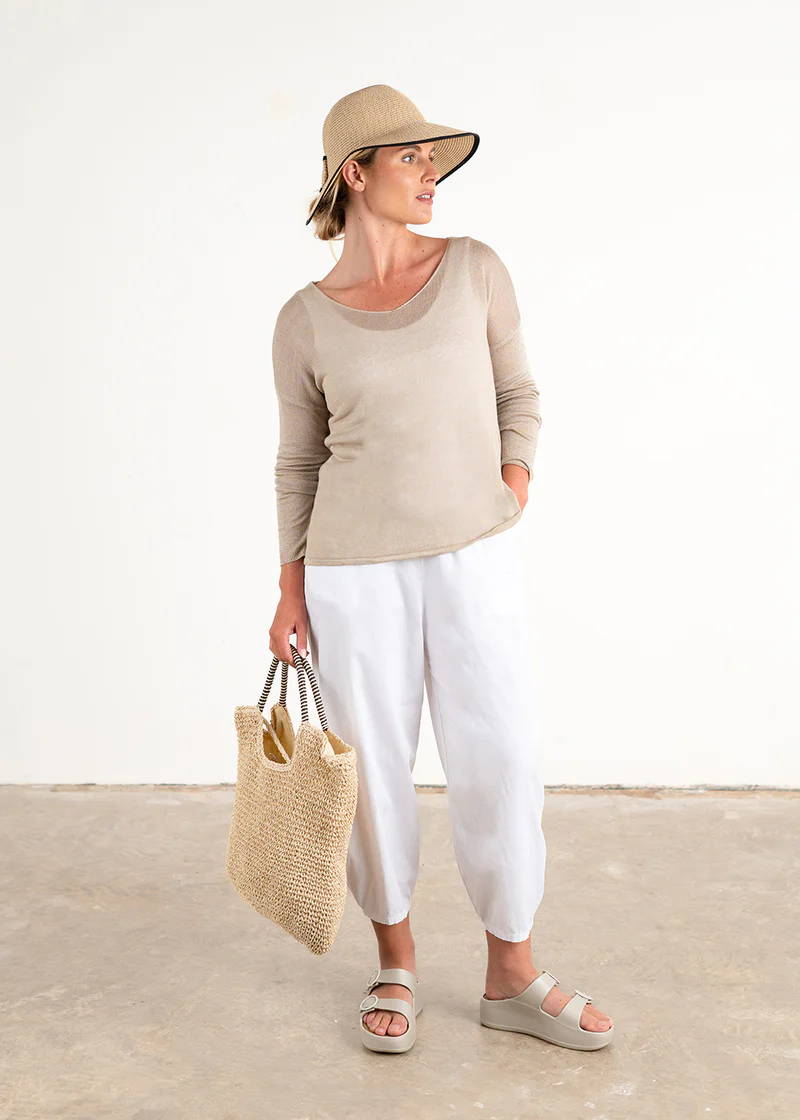 A model wearing a sandy beige semi sheer knitted sweater over a white top, white trousers, off white chunky platform s;ides and wearing a straw hat and holding a straw bag