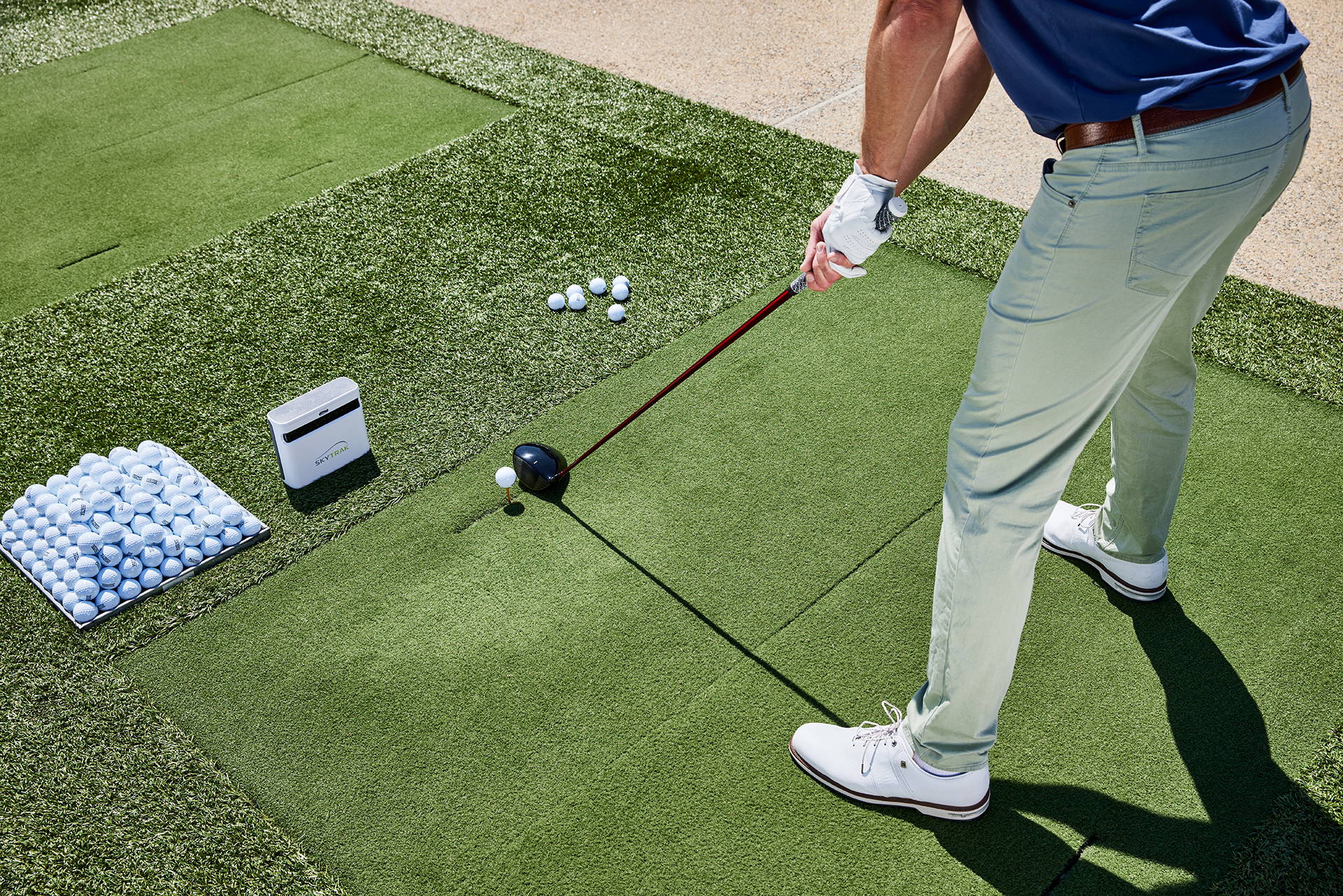 The lower half of a golfer shown while getting ready to swing at a golf range with a SkyTrak+ and a large stack of balls