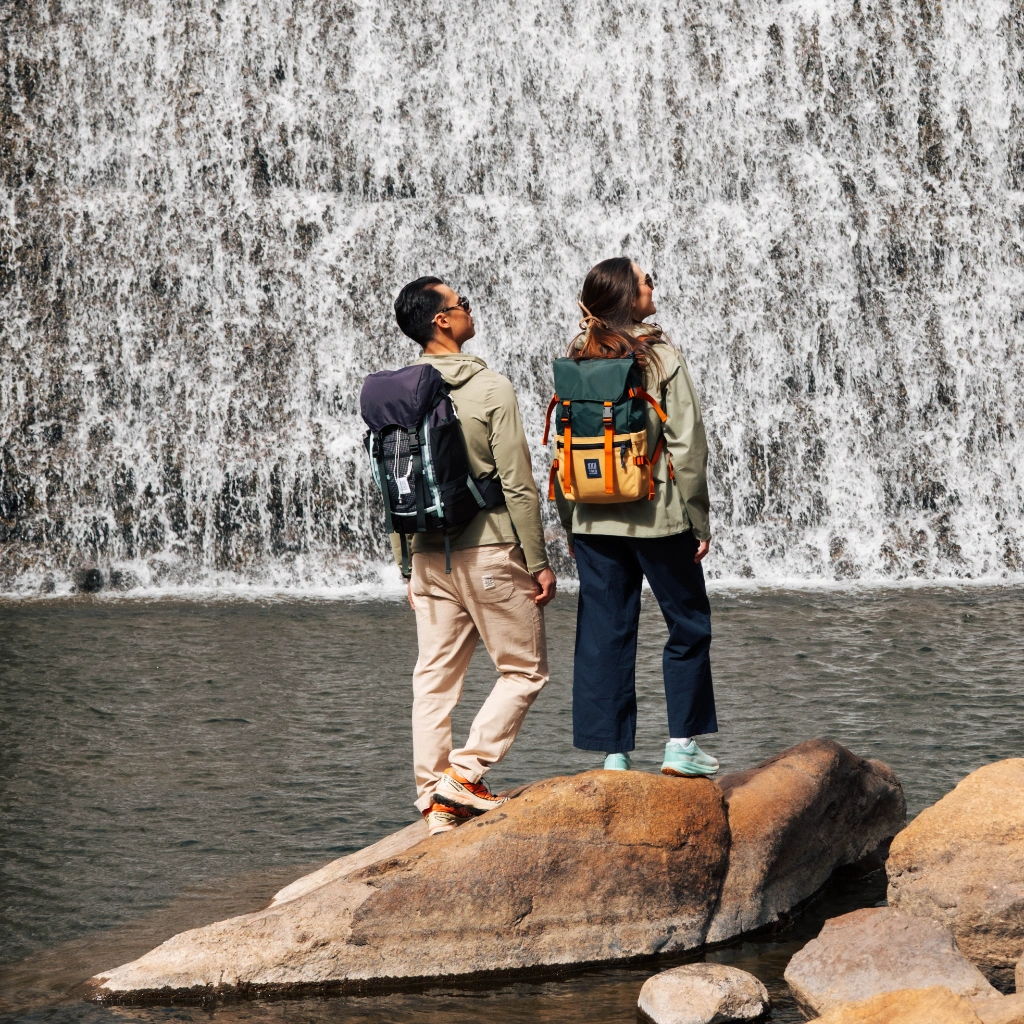 Two people with backpacks standing on rocks near a waterfall.