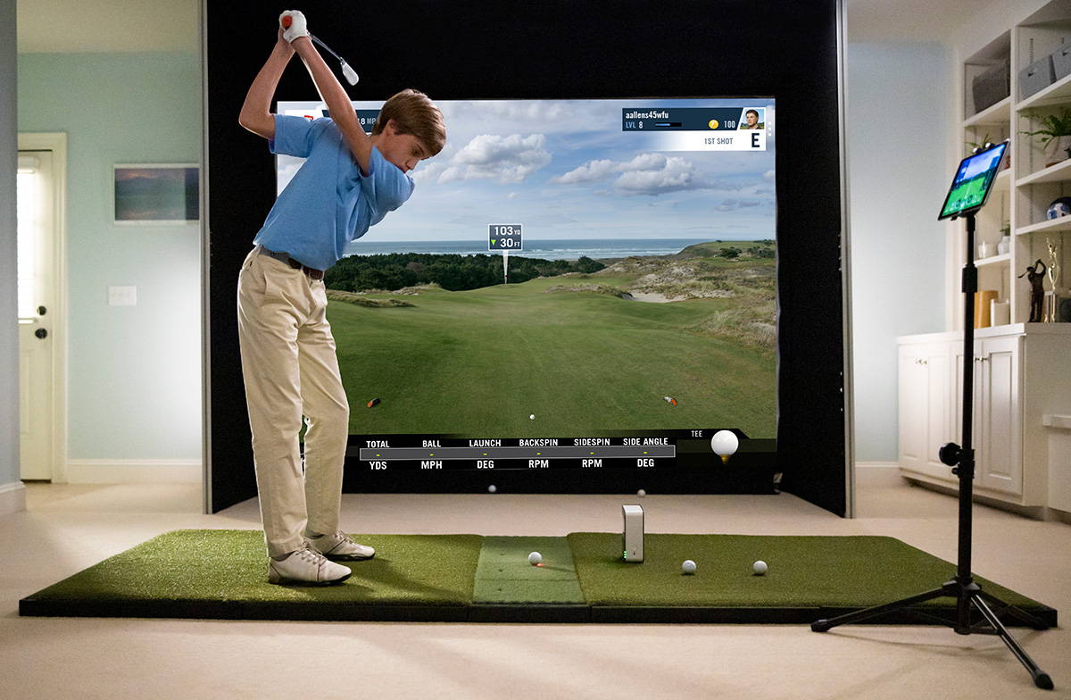 A young man swinging at a golf ball in front of the SkyTrak golf launch monitor into an impact screen with simulation software on it